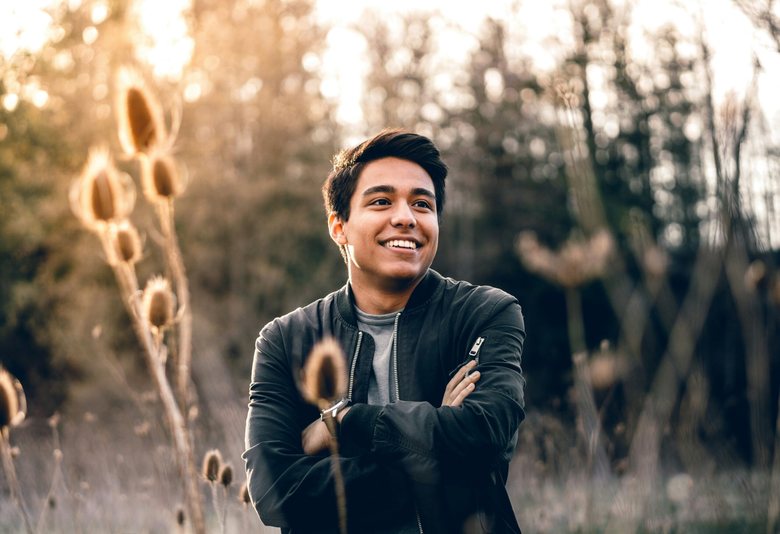 Smiling man standing confidently near lush green trees, wearing a casual outfit, symbolizing positivity and self-assurance.